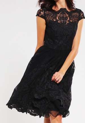 Mujer con un vestido negro de encaje con patrones florales, mangas cortas y cintura ajustada, sosteniendo suavemente la falda mientras está de pie frente a un fondo blanco.