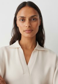 Beige top met lange mouwen, een geribde kraag en V-halsontwerp, met een gladde textuur en een ontspannen pasvorm. Subtiele stiksel details nabij de halslijn.