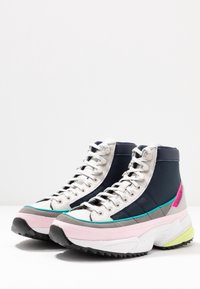 Vyšší tenisky v tmavě modré, bílé a světle růžové barvě. Mají texturované materiály, silné podrážky a barevné akcenty v tyrkysové a zelené.