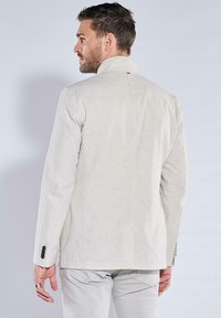 Helles beiges Blazer, gewebte Textur, taillierte Passform, Ein-Knopf-Verschluss, schmale Revers, schwarze Akzentknöpfe, über hellen Hosen getragen.