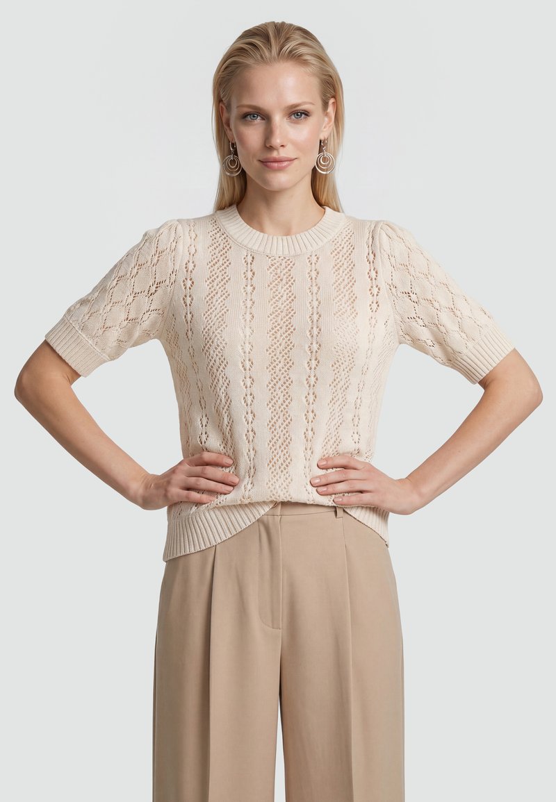 Femme aux cheveux blonds porte un haut en maille crème à manches courtes avec un détail en dentelle et un pantalon beige taille haute à jambes larges, mains posées sur les hanches.