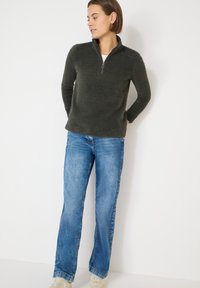Tmavě zelený pulovr se zapínáním na zip u krku z měkké látky, kombinovaný se světle modrými džíny. Pulovr má volný střih a dlouhé rukávy.