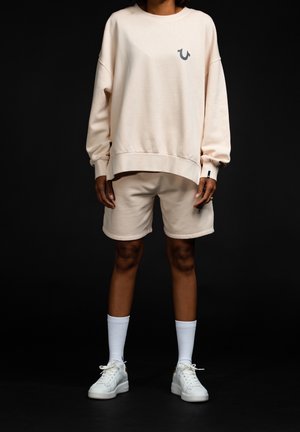 Béžová mikina se šedým logem, doplněná o odpovídající šortky. Bílé tenisky a ponožky dokončují outfit. Měkká textura, ležérní střih.