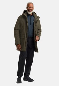 Parka vert olive avec capuche, dotée de plusieurs poches et d'une fermeture éclair. Associée à un haut bleu et un pantalon noir, avec des chaussures noires.