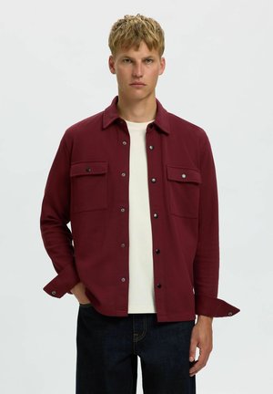 Jeune homme aux cheveux blonds courts portant une chemise bordeaux boutonnée par-dessus un t-shirt blanc et un jean foncé sur un fond clair uni.