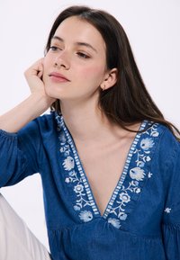 Denimbluse mit tiefem V-Ausschnitt, verziert mit blauer floralem Stickerei und gestreiften Akzenten entlang des Ausschnitts; lange Ärmel mit gerafften Manschetten.