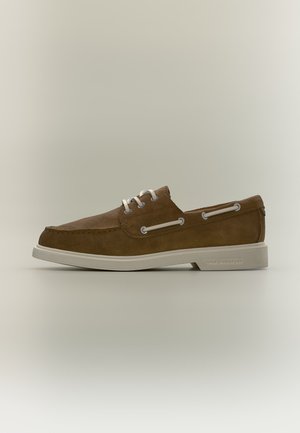 Zapato náutico de ante marrón con cordones y suela blancos, con ojales metálicos y detalle de cordones lateral, presentado sobre un fondo neutro.