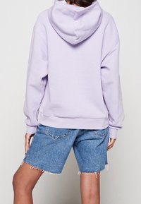 Persona che indossa un cappuccio lilla chiaro e pantaloni denim blu con orli sfrangiati, in piedi con la schiena rivolta verso uno sfondo semplice.