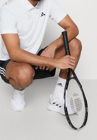 Polo blanc avec logo Adidas noir, short noir, baskets blanches, tenant une raquette de tennis noire avec une prise texturée.