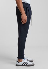 Navy krāsas tapered joggeri ar teksturētu audumu, elastīgu jostasvietu, ar aukliņu, kā arī sānu kabatām, apakšā kombinēti ar baltu un pelēku sporta apavu pāri.