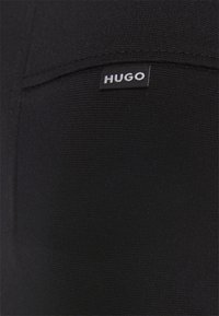 Tela negra texturizada que presenta una pequeña etiqueta negra con texto en blanco que dice "HUGO". El material muestra un patrón tejido elegante.