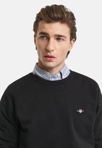 Black sweatshirt with a crew neck, featuring a small embroidered logo on the chest. Worn over a light blue striped collared shirt.