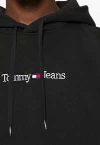 Sudadera negra con el texto "Tommy Jeans" y el logotipo de la bandera rectangular en rojo, blanco y azul bordado en el pecho, usada por una persona.