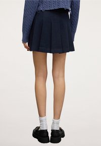 Námořnická sukně s plisováním, vyrobená z látky, s přiléhavým pasem a rozšířeným spodním lemem. Spárovaná s černými botami a bílými, žebrovanými ponožkami.