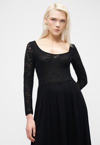 Γυναίκα με ξανθά μαλλιά, που φοράει μακρυμάνικο μαύρο δαντελένιο φόρεμα με λαιμόκοψη scoop, που στέκεται μπροστά σε απλό φωτεινό φόντο.