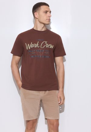 Uomo con maglietta marrone "Work Crew" e pantaloncini beige in piedi davanti a uno sfondo bianco, guarda a destra con le mani nelle tasche.