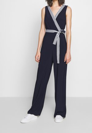 Kobieta w granatowym, bezrękawnym kombinezonie z białym paskiem w paski zawiązanym wokół talii i białymi sneakersami, stojąca na tle jasnego, jednolitego tła.