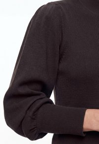 Gros plan d'une personne portant un pull en maille côtelée foncée avec des manches bouffantes resserrées au poignet.