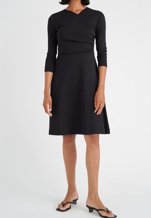 Czarna sukienka z dopasowaną, kopertową górą, rękawami do połowy przedramienia oraz rozkloszowanym dołem. Gładka faktura i długość do kolan.