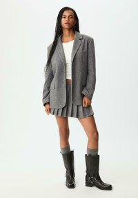 Blazer grigio in lana, top corto bianco, gonna plissettata grigia e stivali neri in pelle al ginocchio con fibbie. Texture morbida, vestibilità sartoriale e look a strati.