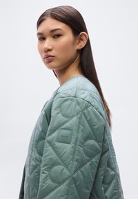 Ανοιχτό πράσινο μπουφάν με επένδυση, χαρακτηριστικό ραφή διαμαντιού, κυκλικά διακοσμητικά στοιχεία και τετράγωνη ετικέτα της μάρκας στον αριστερό ώμο. Ομαλή υφή.