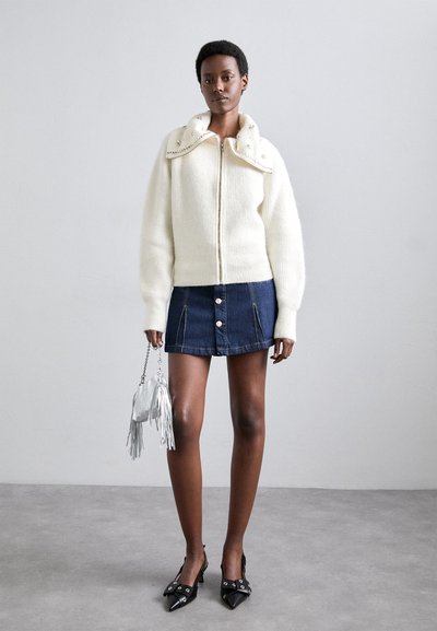 White knitted zip-up sweater with a structured collar, paired with a blue denim mini skirt and black pointed-toe heels. Holding a silver fringed bag.