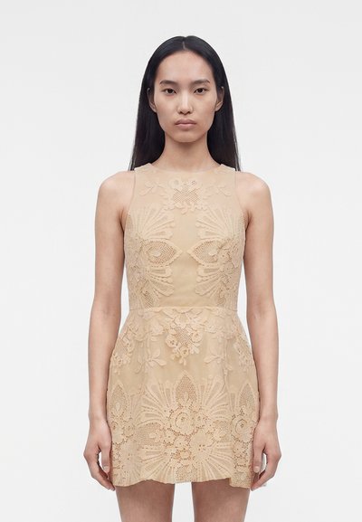 Femme aux longs cheveux noirs portant une robe en dentelle beige sans manches avec des motifs floraux, debout devant un fond blanc uni.