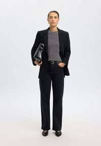 Črni krojeni blazer čez siv pleteni pulover, kombiniran s črnimi širokimi kavbojkami in črnimi ostrimi balerinkami, drži črno torbico.