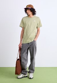 Oliven grøn t-shirt med et hvidt logo, grå jeans, hvide og sorte sneakers, brun kasket med farvet logo, bærende en brun tote bag.