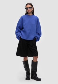Übergroßer blauer Strickpullover mit rundem Ausschnitt, kombiniert mit schwarzen, gefalteten Shorts und kniehohen schwarzen Lederstiefeln mit Schnallen.