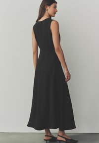 Musta hihaton mekko, jossa on tiukka yläosa, levenevä helma ja takana vetoketjukiinnitys. Mallilla on päällään mustat slingback-kengät. Suora kangasintenssity.