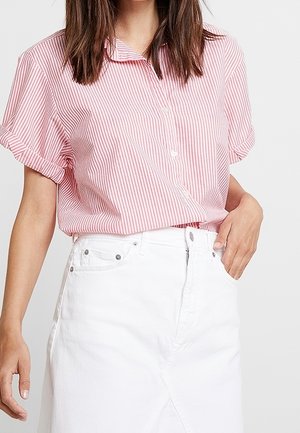 Woman wearing a pink and white striped short-sleeve shirt tucked into a high-waisted white skirt, with one hand in pocket.