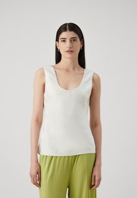 Weißes ärmelloses Oberteil mit einem runden Ausschnitt, aus glattem Stoff gefertigt. Kombiniert mit plissierten, hellgrünen weiten Hosen.