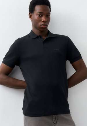 Νέος άνδρας που φοράει μαύρο πόλο με κοντά μανίκια και το λογότυπο «BOSS», στέκεται με τα χέρια στην πλάτη, μπροστά σε λευκό φόντο.