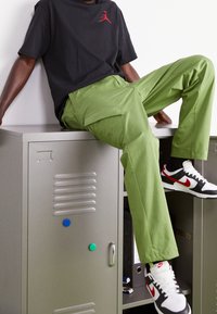 Groene cargobroek, zwart T-shirt met rood logo, en zwart/rode sneakers. Persoon zit op een grijze locker, met kleurrijke magneten erop bevestigd.