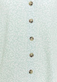 Light green shirt with a subtle leaf pattern, featuring five round brown buttons down the front and a soft fabric texture.