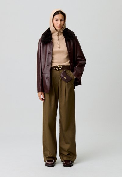 Veste en cuir marron avec col en fourrure noire, sweat à capuche beige en tricot, pull crème, pantalon large olive et chaussures marron avec des coutures blanches.