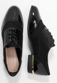 Des chaussures oxford noires en association de cuir verni et de suède. Détails avec des perforations et une accentuation dorée sur le talon.