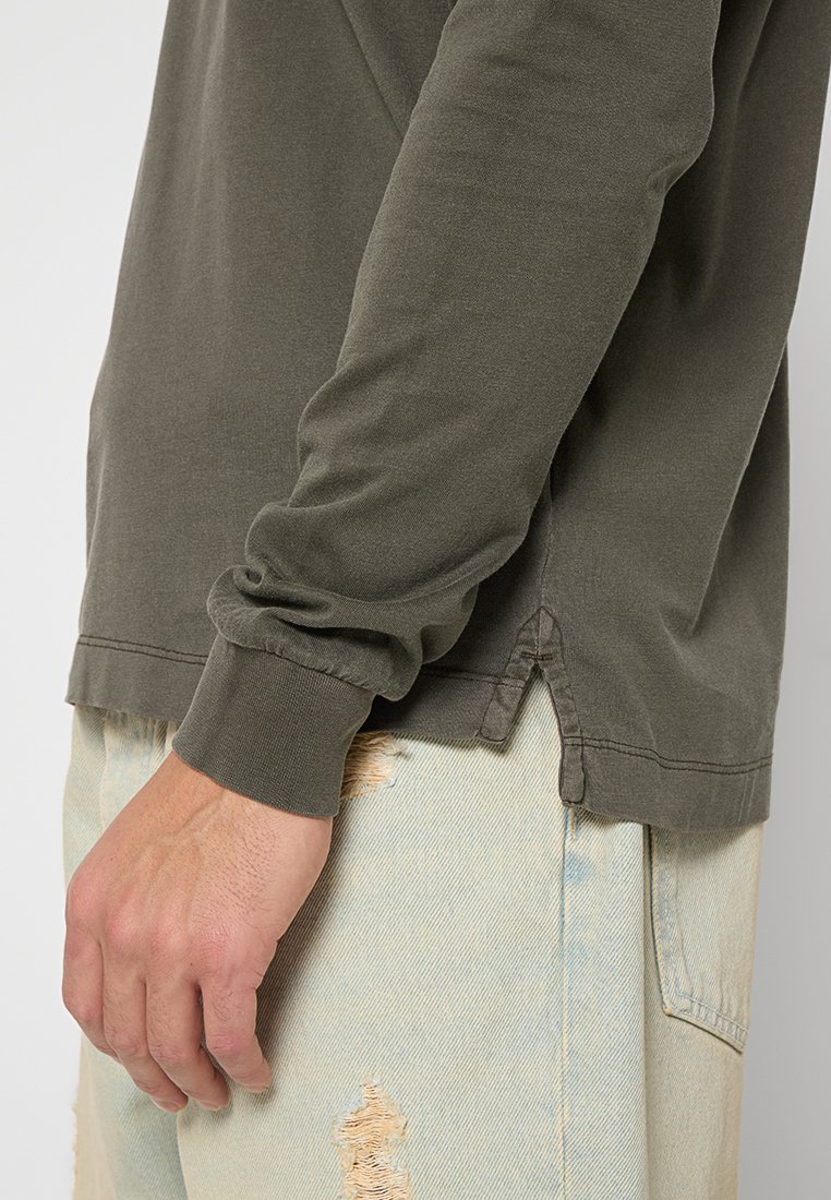 Olivengrünes Langarmshirt mit Rundhalsausschnitt, ausgestattet mit gerippten Bündchen und seitlichen Schlitzen, kombiniert mit hellen, ausgefransten Jeans.