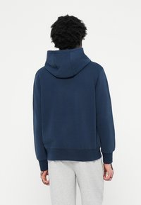 Uomo con capelli ricci che indossa una felpa blu e pantaloni da jogging grigi chiari, in piedi di spalle contro uno sfondo bianco uniforme.