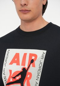 Μαύρο βαμβακερό μπλουζάκι με έντονα κόκκινα και λευκά γραφ ικά που απεικονίζουν το κείμενο "AIR JORDAN" και μια σιλουέτα παίκτη σε δράση.