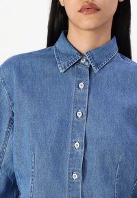 Camicia in denim azzurro chiaro con colletto a punta, bottoni frontali e una texture liscia; presenta due tasche laterali e una vestibilità rilassata.