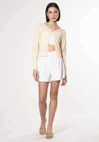 Cardigan color crema con scollatura rotonda e un bottone centrale, abbinato a pantaloni corti bianchi a vita alta. Tessuto leggero e testurizzato.