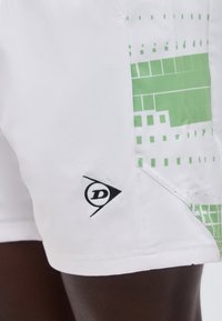 Fehér rövidnadrág zöld geometriai mintával az oldalán és egy kis fekete logóval a szegély közelében, sötét bőrön viselve.