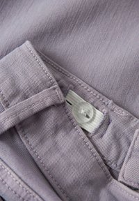 Violeta auduma, ar gludu tekstūru, ar pogu aizdari un izšūtu detaļu pa malu. Ietver baltu etiķeti ar tekstu.