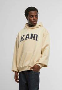 Beige oversized hoodie i mjukt tyg, med en framficka och "KANI" tryckt i marinblå bokstäver över bröstet.
