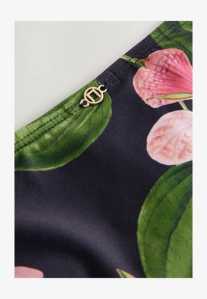 Kukallinen kangas, jossa on suuria vaaleanpunaisia kukkia ja vihreitä lehtiä tummalla taustalla; lähellä saumaa on kultainen pyöreä koriste.