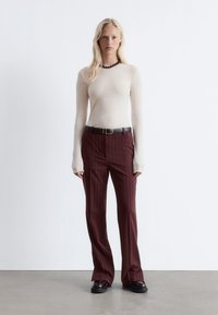 Beige Langarmshirt, figurbetont, mit weiten Manschetten; burgunderfarbene gestreifte Hose, mit geradem Schnitt, kombiniert mit schwarzen klobigen Schuhen und Gürtel.