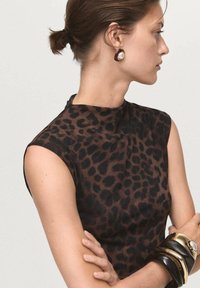 Kvinna i ärmlös mörkbrun leopardmönstrad topp, bär stora guld- och svarta armband samt en brun örhänge med en vit pärldetalj, håret i en knut.