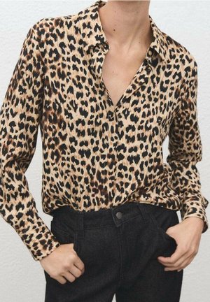 Personne portant un chemisier à manches longues imprimé léopard, rentré dans un pantalon noir taille haute, les mains dans les poches.